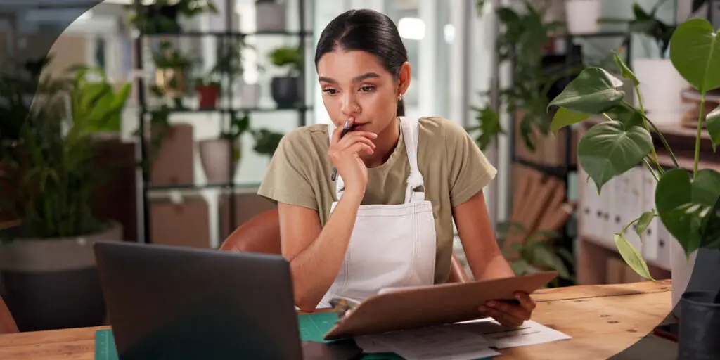 Woman surrounded by potted plants looks at a clipboard and laptop screen