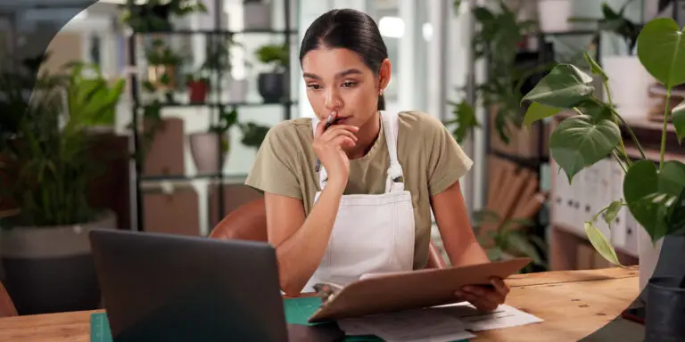 Woman surrounded by potted plants looks at a clipboard and laptop screen